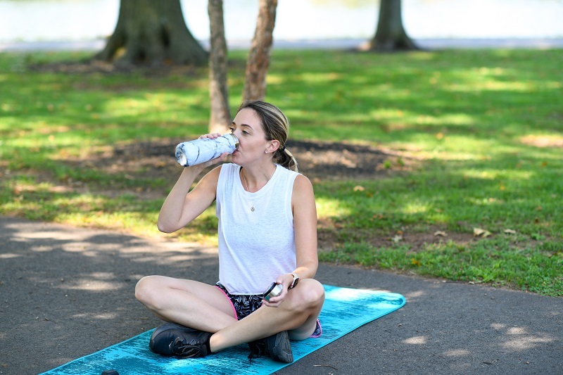 Woman in athletic clothes, sitting on a yoga mat, outside, drinking water.